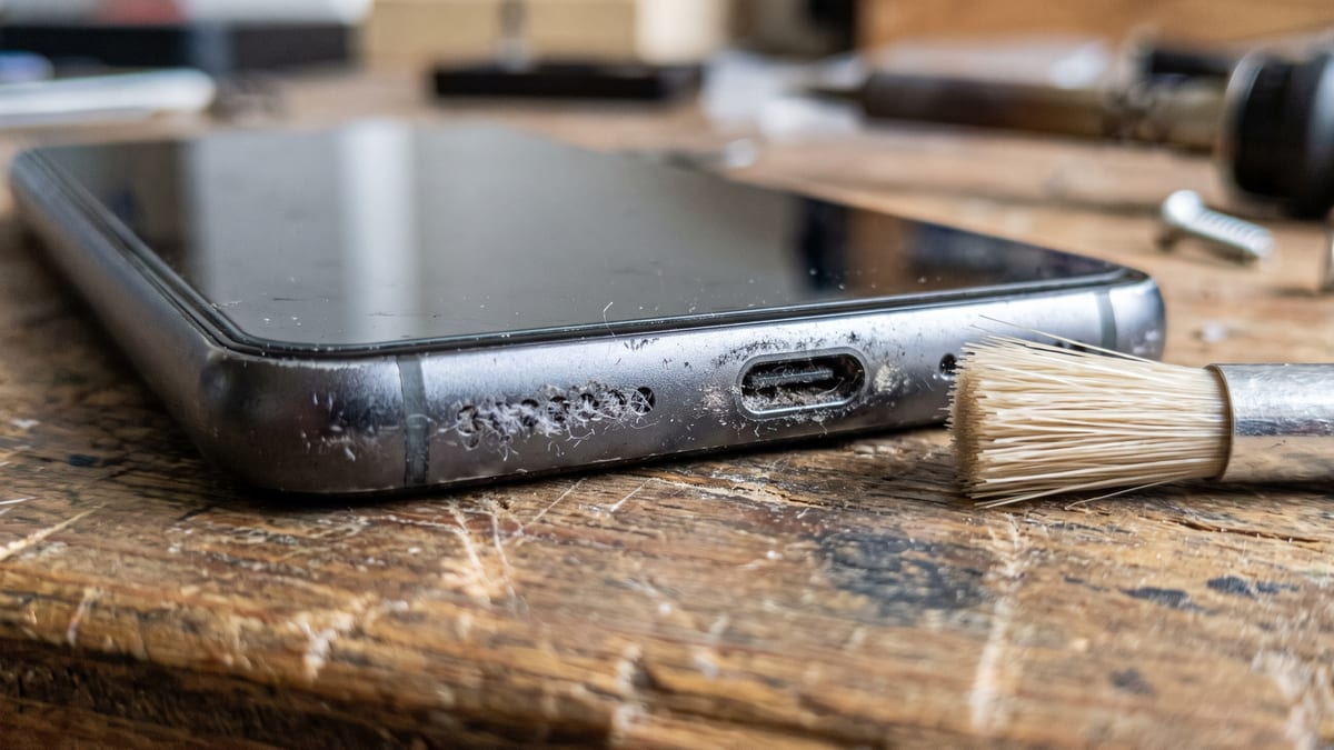 Close da parte de baixo de um celular na bancada, mostrando a grade do alto-falante ao lado de uma escova de cerdas macias